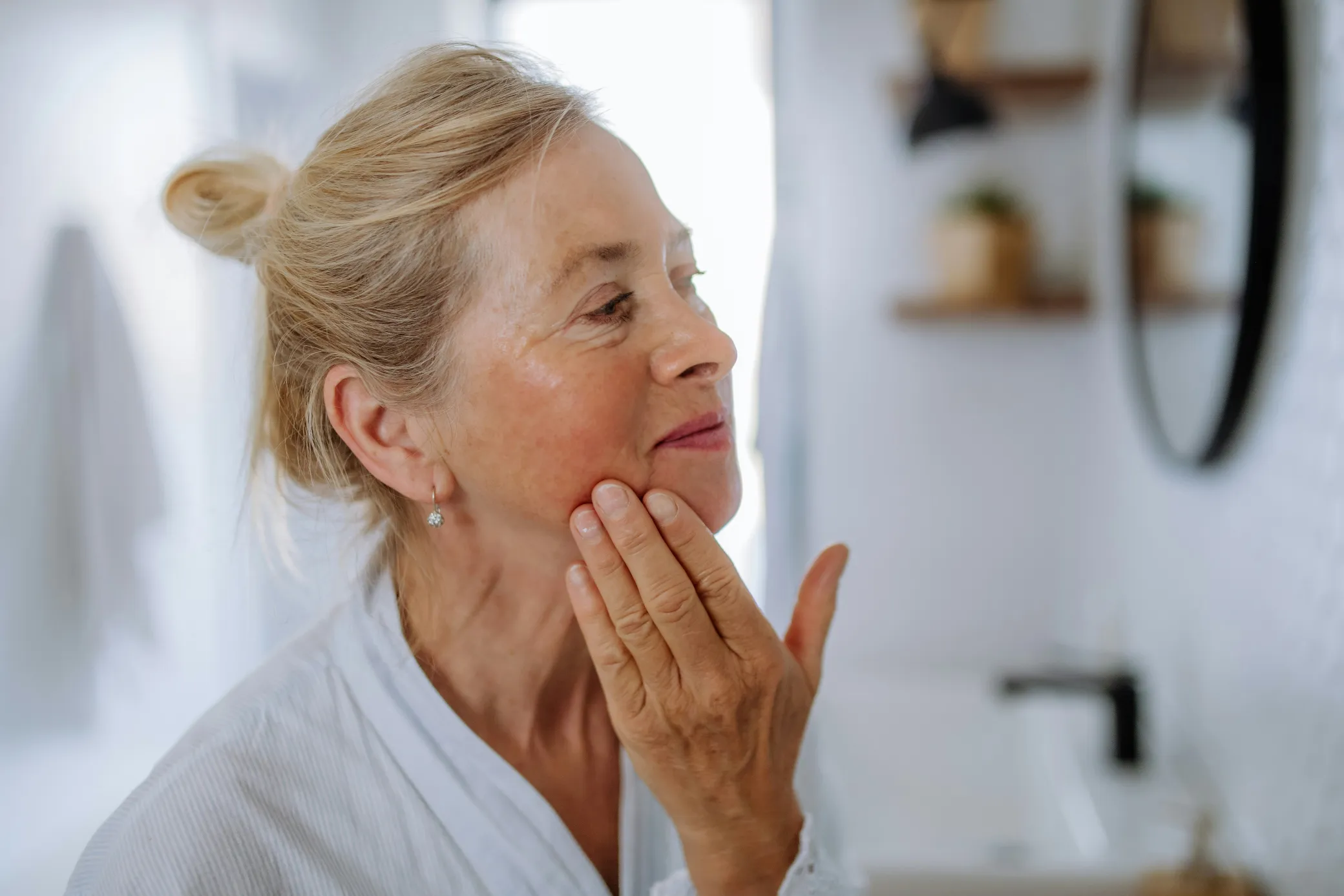 Woman gently touching her face in a serene bathroom setting, reflecting personalized care at a Colorado Springs medical aest…